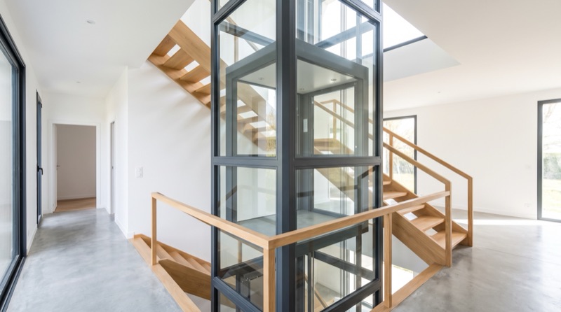 Ascenseur électrique panoramique vitré desservant deux niveaux le long d'un escalier bois